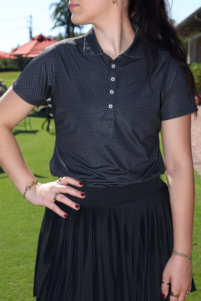Woman in a polka dot shirt and pleated skirt stands with hand on hip. Sunlit outdoor setting with greenery and relaxed ambiance.