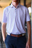 A person is standing indoors, wearing a short-sleeved white polo shirt with a blue and red pattern and navy pants, with their hands in their pockets.