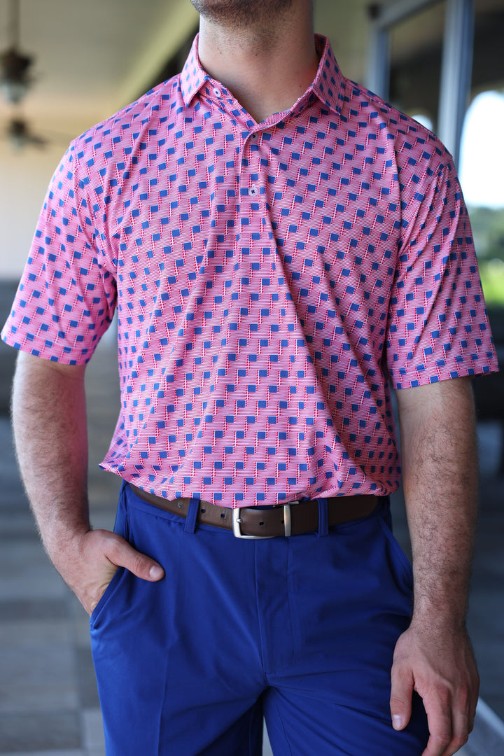 A man is standing with one hand in his pocket, wearing a short-sleeved pink shirt patterned with small flags and blue pants.