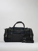 A duffel bag with a green and blue plaid fabric is pictured against a plain background. It features black leather handles and a detachable strap, with silver zippers