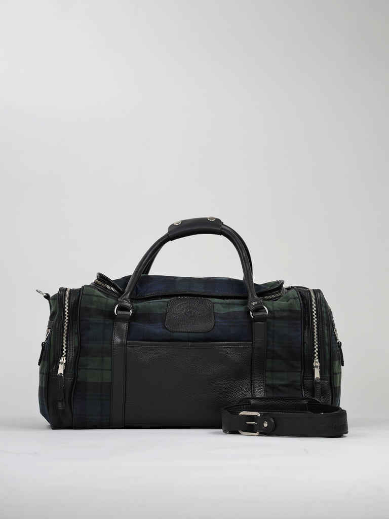 A duffel bag with a green and blue plaid fabric is pictured against a plain background. It features black leather handles and a detachable strap, with silver zippers