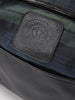Close-up of a black leather bag with embossed logo on a textured patch, set against a green and blue plaid fabric lining