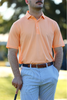 A man wearing an orange polo shirt and striped pants stands outdoors, holding a golf club.