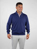 Smiling man wearing a blue half-zip sweater over a checked shirt and light gray pants, standing against a plain white background.