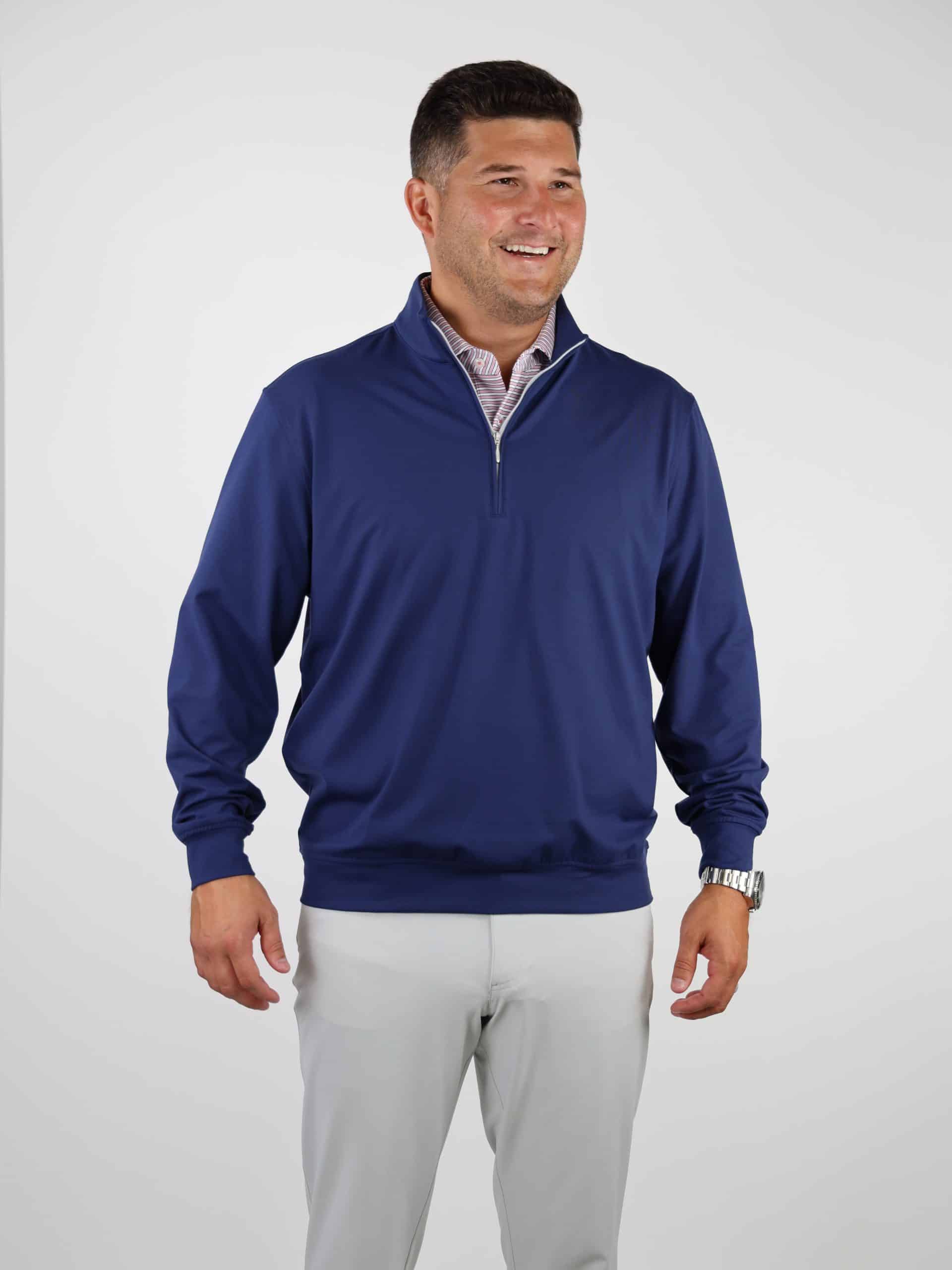 Smiling man wearing a blue half-zip sweater over a checked shirt and light gray pants, standing against a plain white background.
