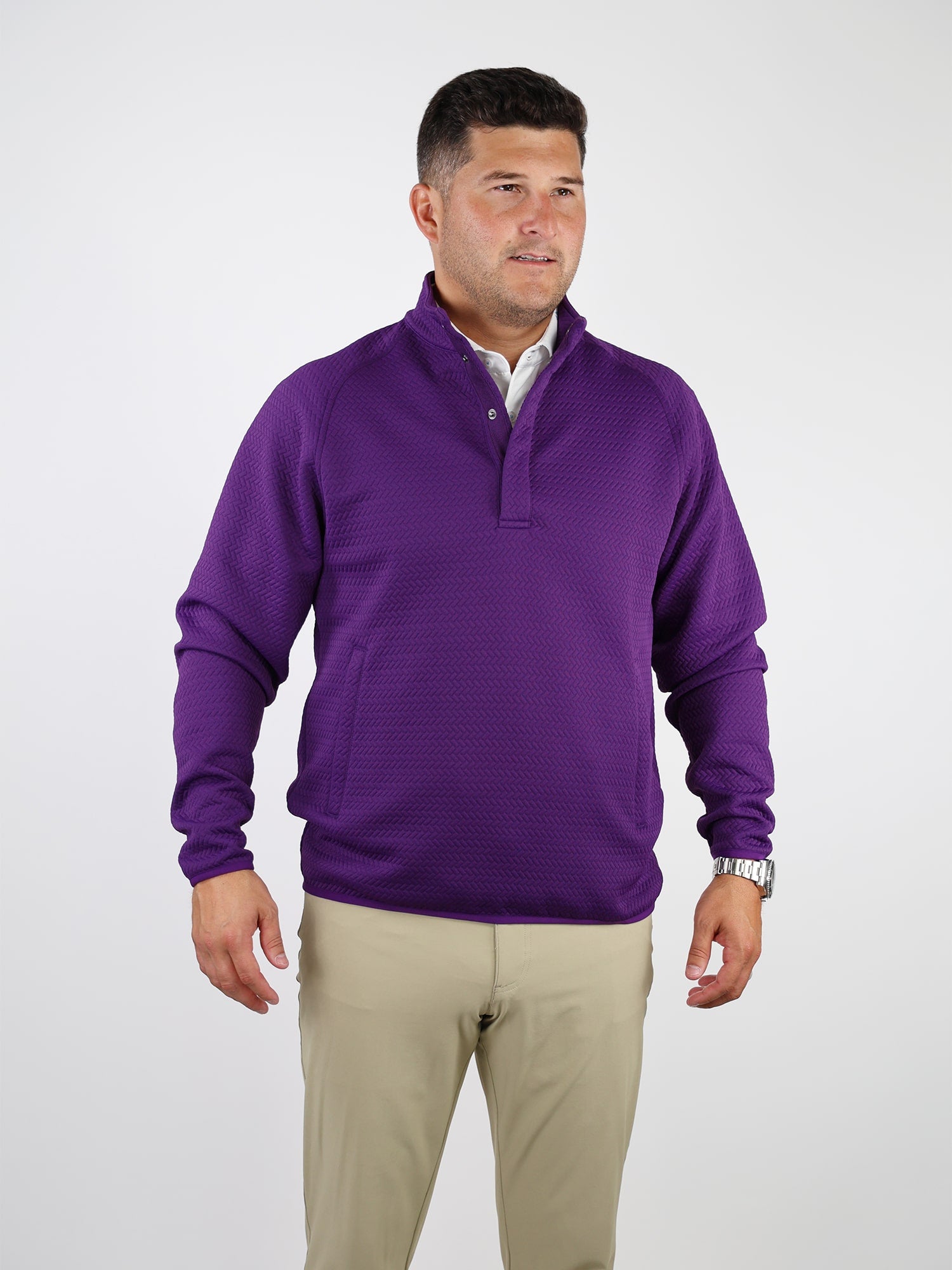 A person wearing a vibrant purple performance golf pullover and beige pants stands against a plain white background, exuding a composed and confident demeanor.