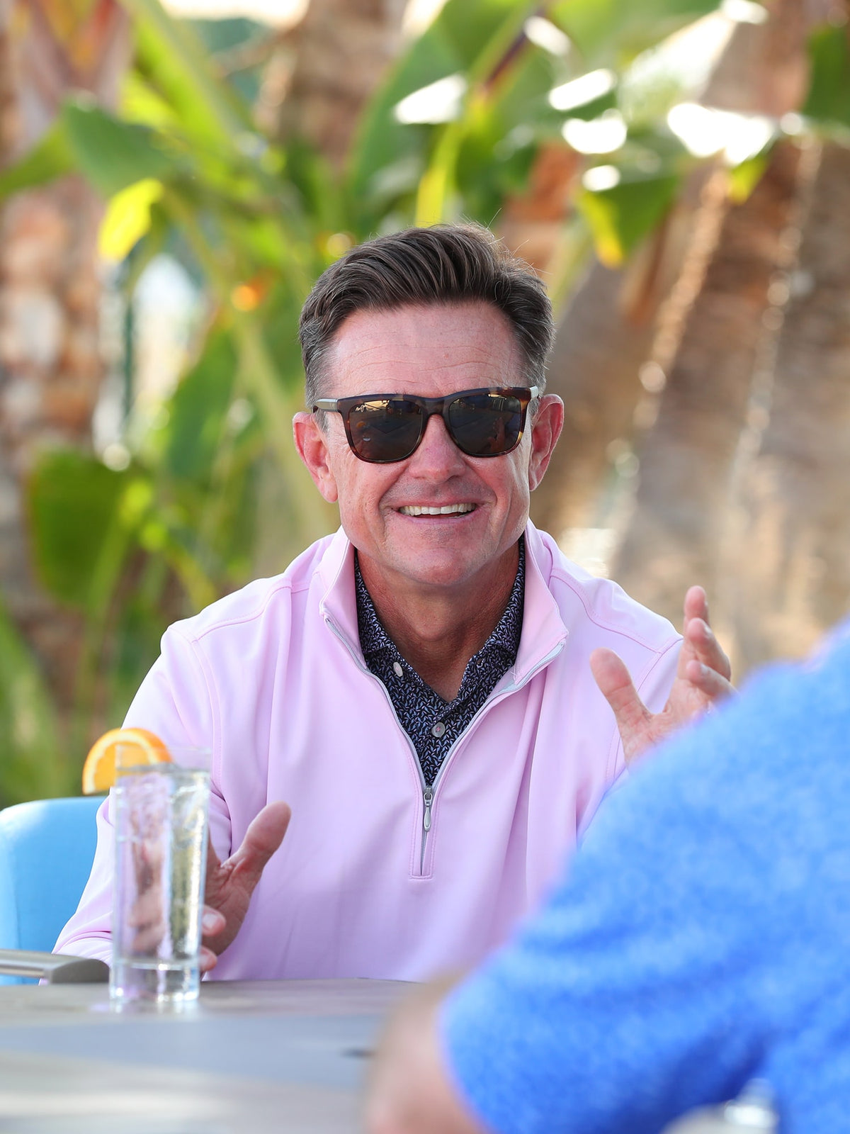 A man in sunglasses and a pink shirt smiles while gesturing at an outdoor table. A drink with an orange slice sits nearby.