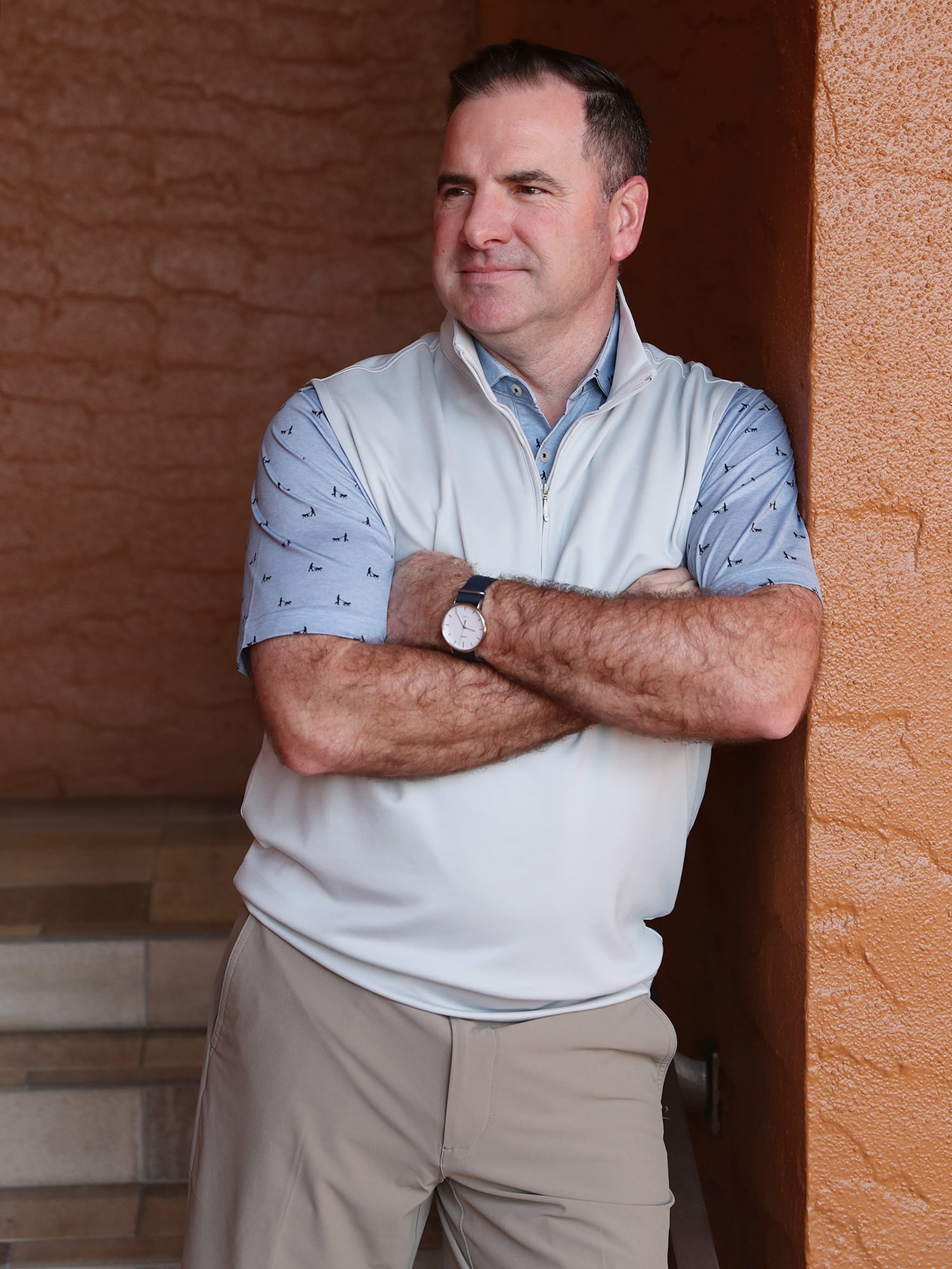 A man with short dark hair leans against an orange wall, arms crossed, wearing a light vest over a patterned shirt.