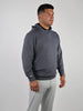 A man in a gray textured hoodie and light-colored pants stands against a plain white background. He gazes to his right with a neutral expression.
