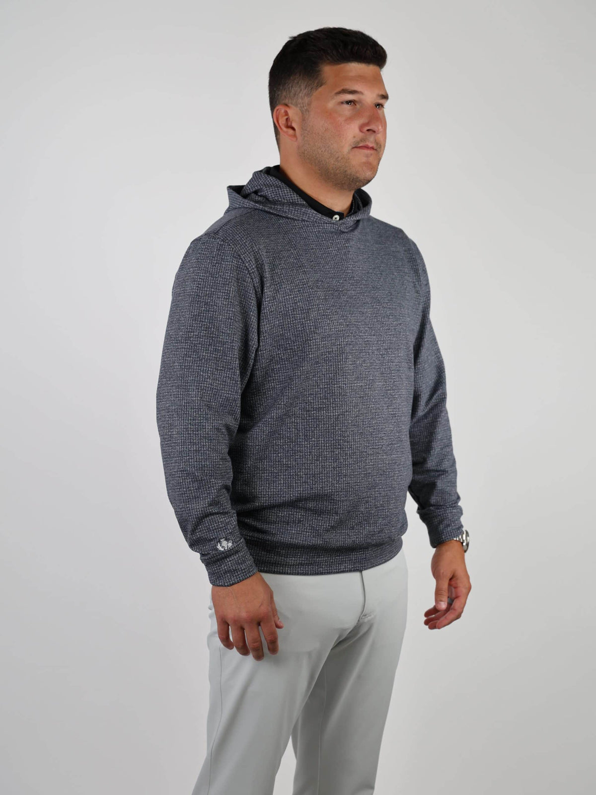 A man in a gray textured hoodie and light-colored pants stands against a plain white background. He gazes to his right with a neutral expression.