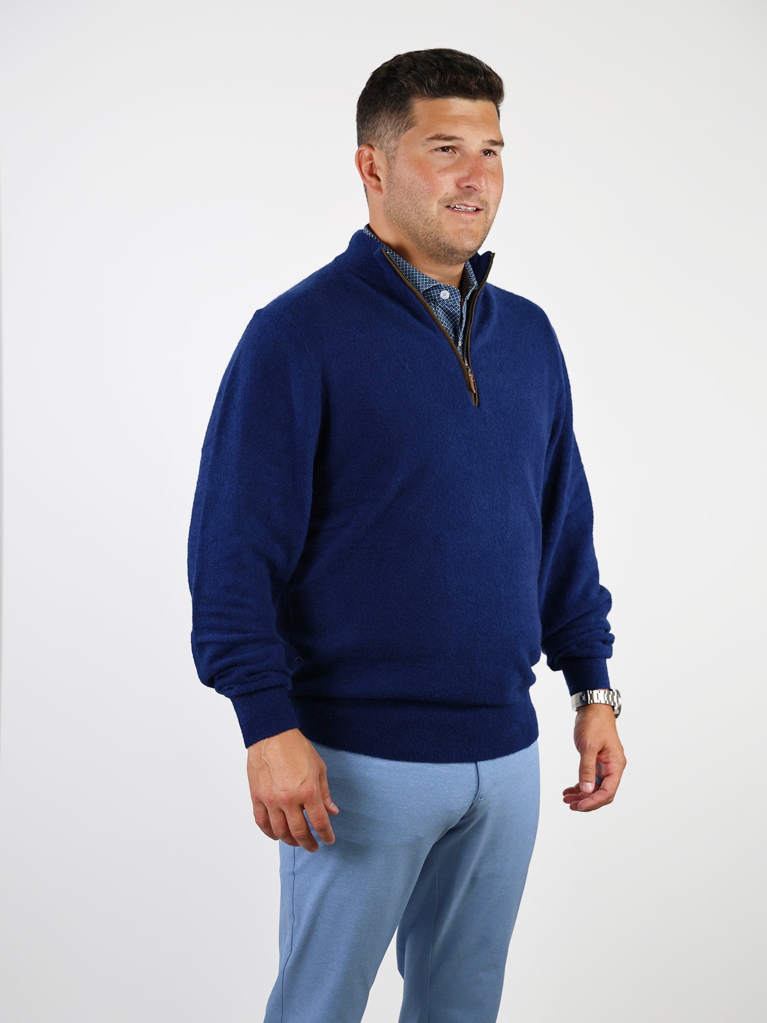 Man wearing a blue quarter-zip sweater over a checkered shirt, paired with light blue pants. He stands against a plain white background, looking confident and relaxed.