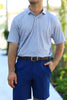 A man stands with his right hand in his pocket, wearing a patterned short-sleeve polo shirt and blue shorts in a greenery-filled outdoor setting.