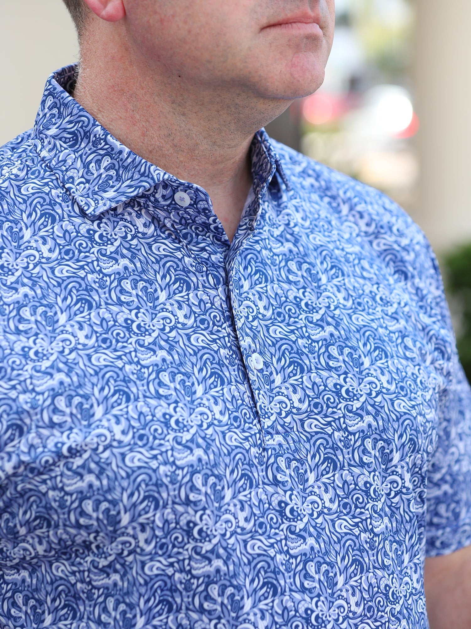 A man wearing a blue and white patterned shirt with intricate floral designs. His expression is neutral, and the background is softly blurred.