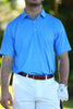 A man wearing a blue patterned polo shirt and white pants stands outdoors with a golf club in his right hand.