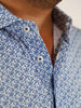 Close-up of a man wearing a blue and white patterned shirt with small floral designs. The shirt is unbuttoned at the top, conveying a casual style.