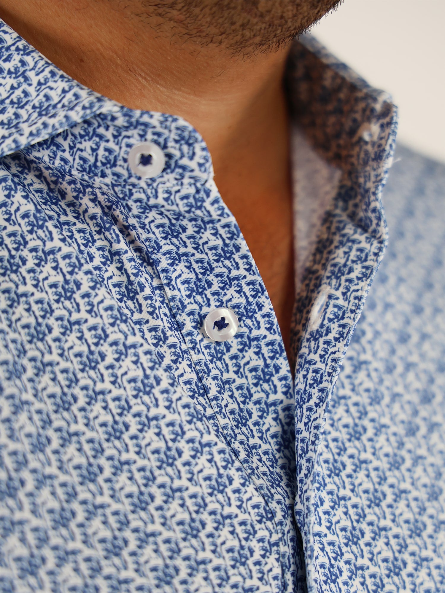 Close-up of a man wearing a blue and white patterned shirt with small floral designs. The shirt is unbuttoned at the top, conveying a casual style.
