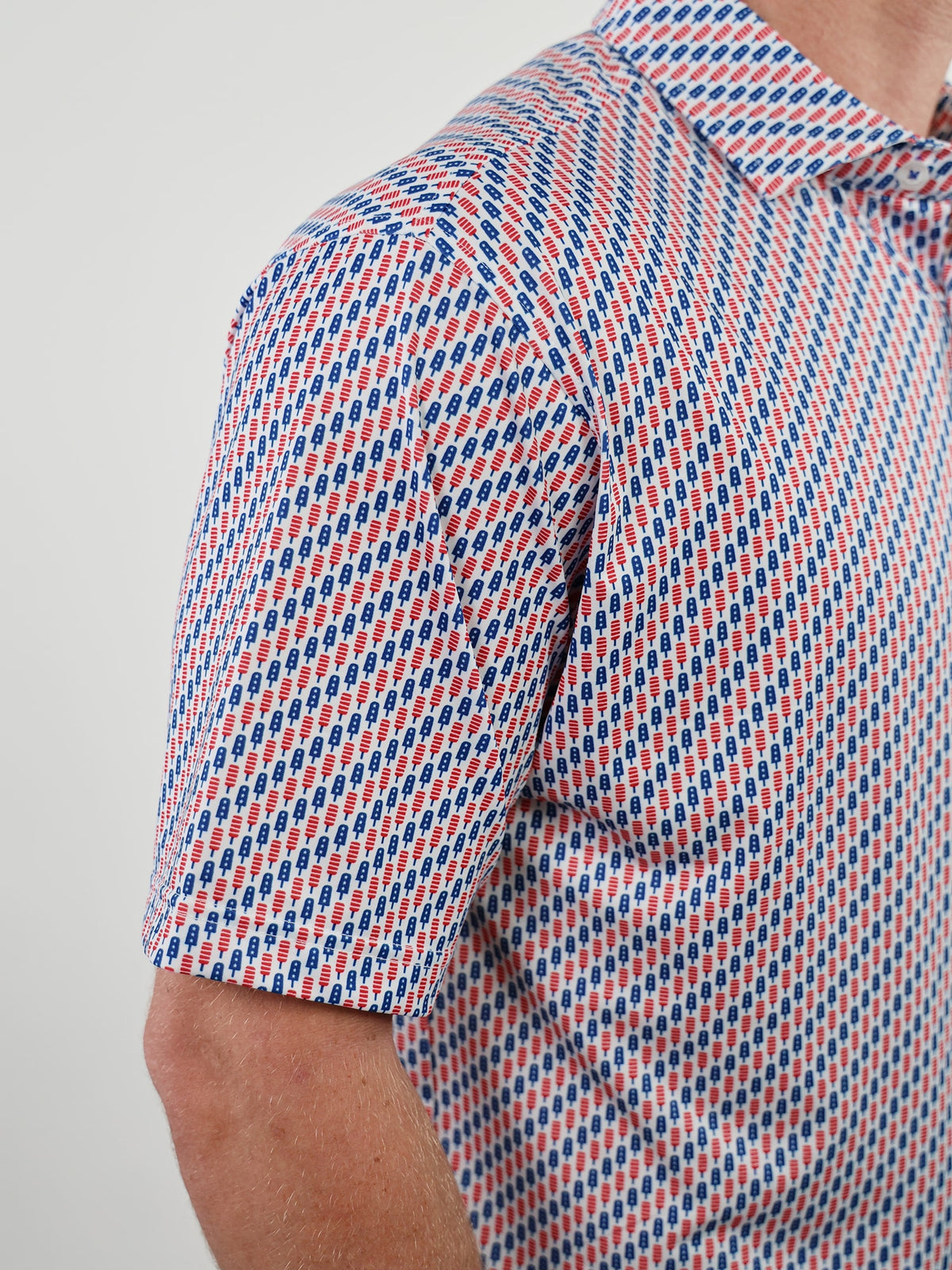 Close-up of a man's short-sleeve shirt with a repeating popsicle pattern in blue and red on a white background. The design is vibrant and modern.