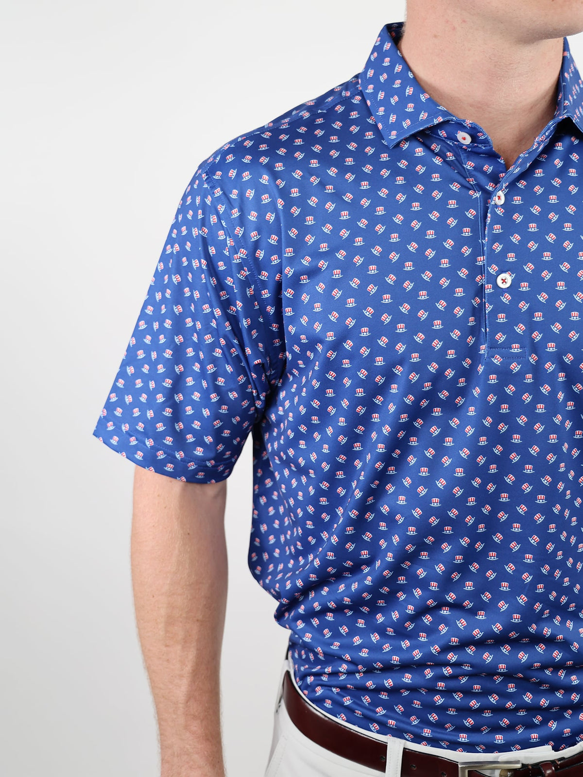 A person wearing a blue golf shirt with a pattern of small Uncle Sam hats. The shirt is tucked into white pants with a brown belt.