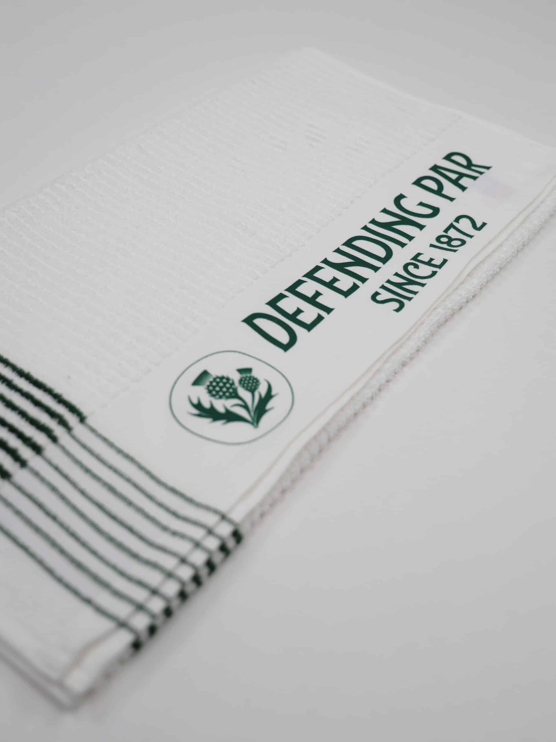 A neatly folded white towel features bold green text reading "Defending Par Since 1872" on a stripe, with a thistle emblem