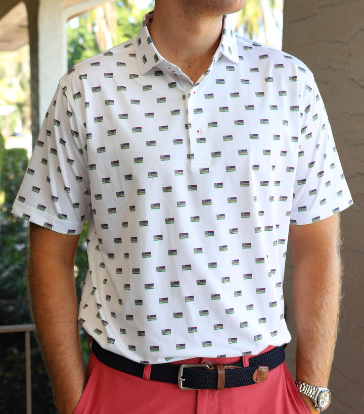 A person wearing a white polo shirt with a pattern of small flags and red pants stands against a blurred outdoor background.