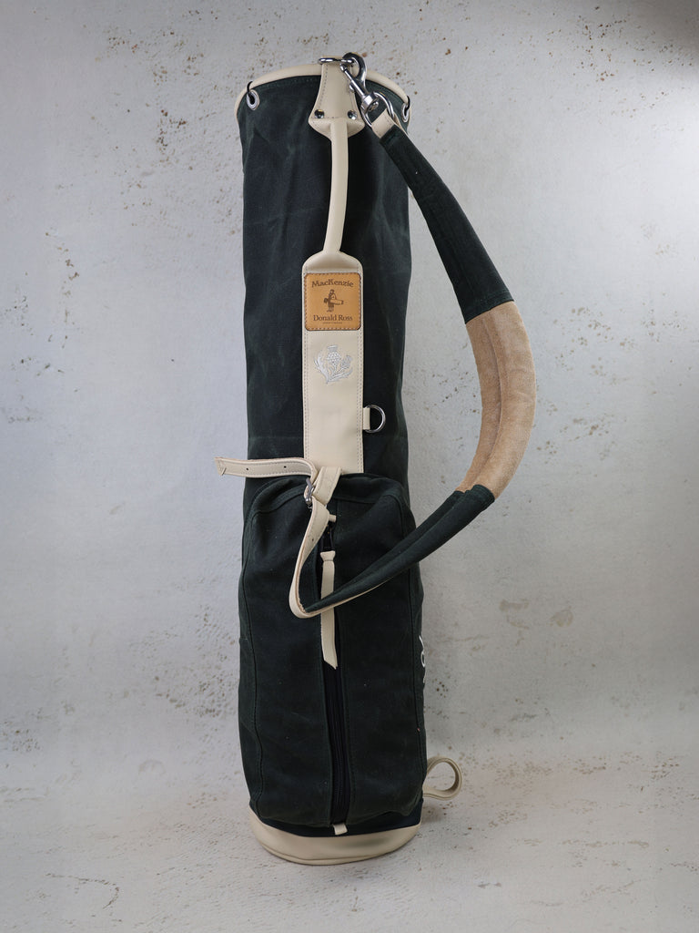 Black and cream canvas golf bag with leather accents and shoulder strap displayed against a concrete backdrop.