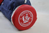 A red circular golf club head cover displaying the Donald Ross Dornoch, U.K. emblem with thistle design and 1872 founding date sits on a concrete surface next to navy blue fabric.