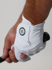 Close-up of a hand wearing a white golf glove gripping a club. The glove features a logo with green and blue accents, conveying a sporty tone.