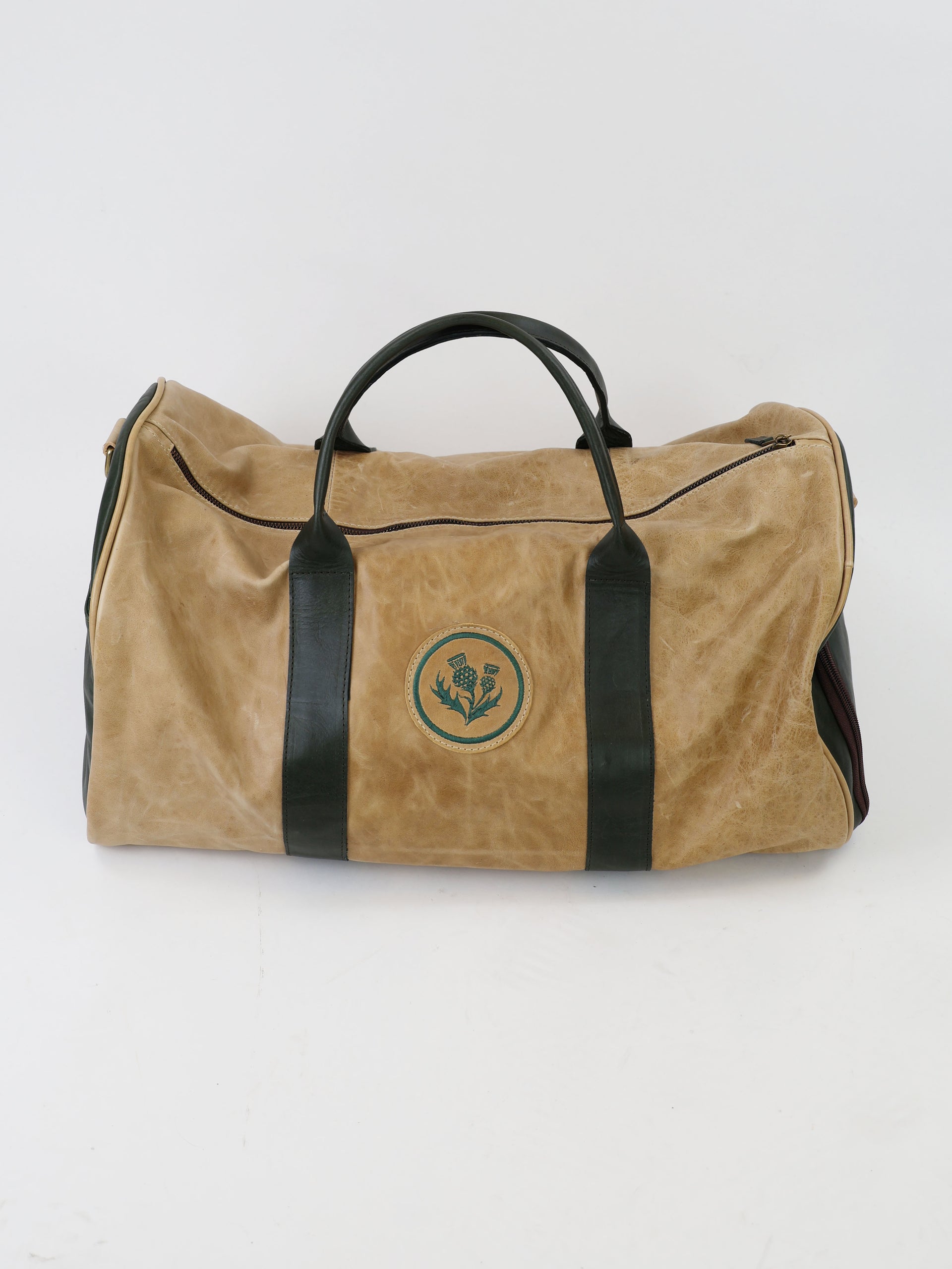 Tan duffel bag with dark handles and a round green emblem featuring floral design. The bag rests on a plain white surface, offering a vintage feel.