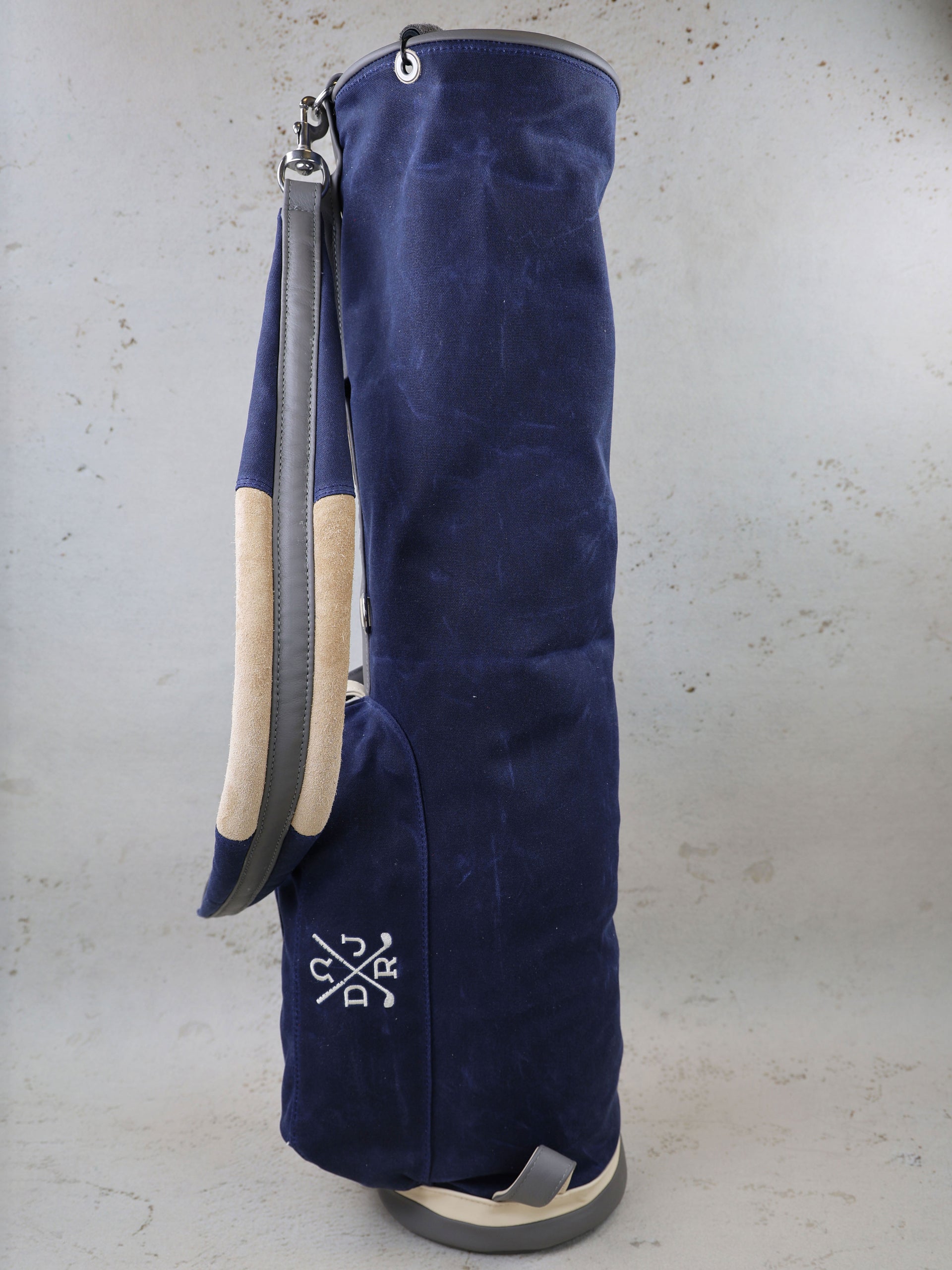 Navy canvas golf bag with cream-colored handles and leather accents, displayed against a concrete background.
