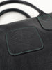 Close-up of a black leather bag with textured grain. Features a stitched label embossed with text and a logo, highlighted with green stitching