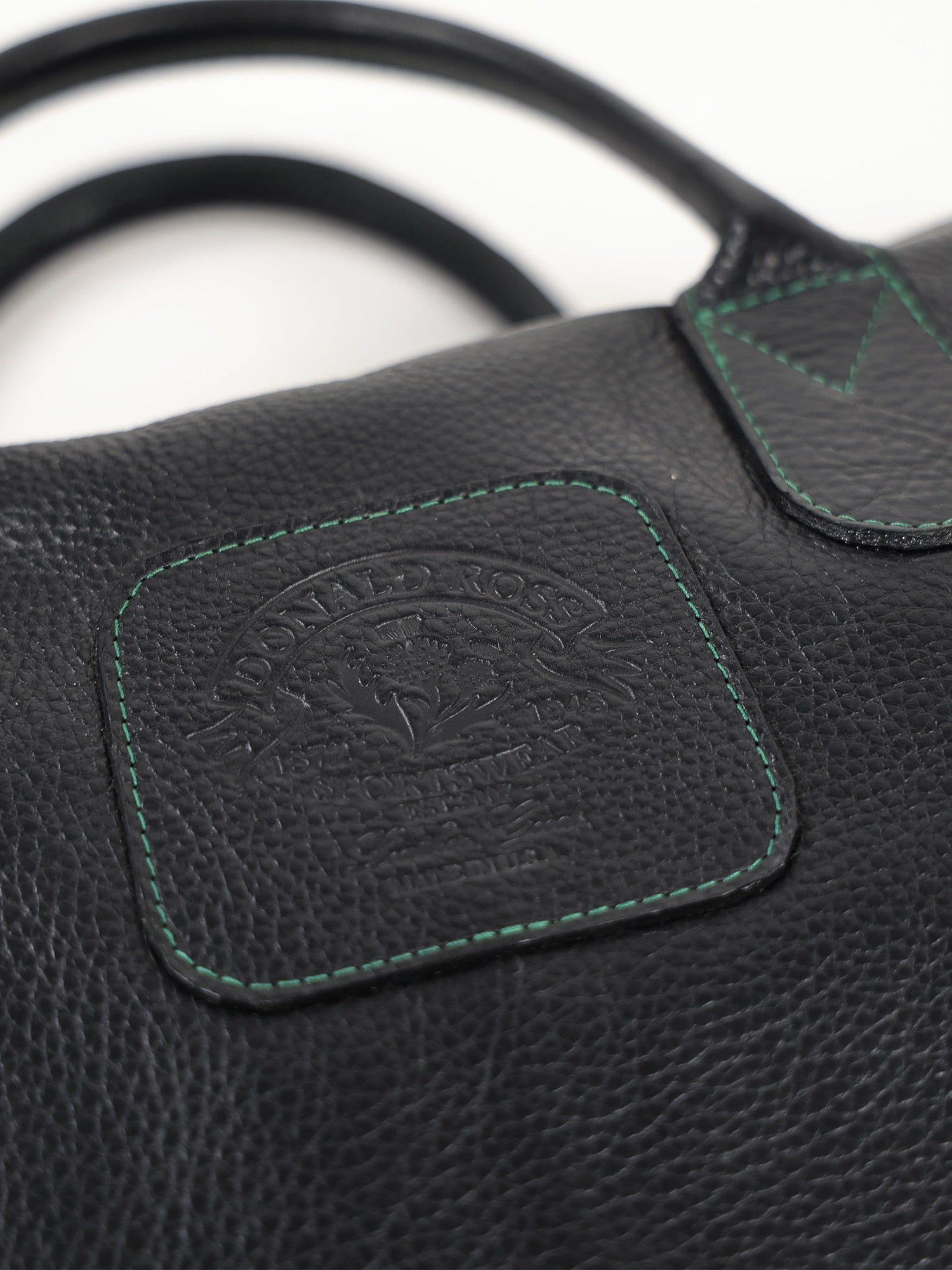 Close-up of a black leather bag with textured grain. Features a stitched label embossed with text and a logo, highlighted with green stitching