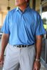 A man stands with his hands at his sides, wearing a blue patterned short-sleeve shirt and light-colored pants.