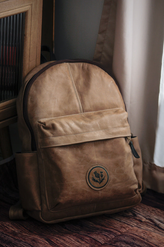 A handcrafted tan leather backpack with a pebble grain finish featuring adjustable shoulder straps and a top carry handle.