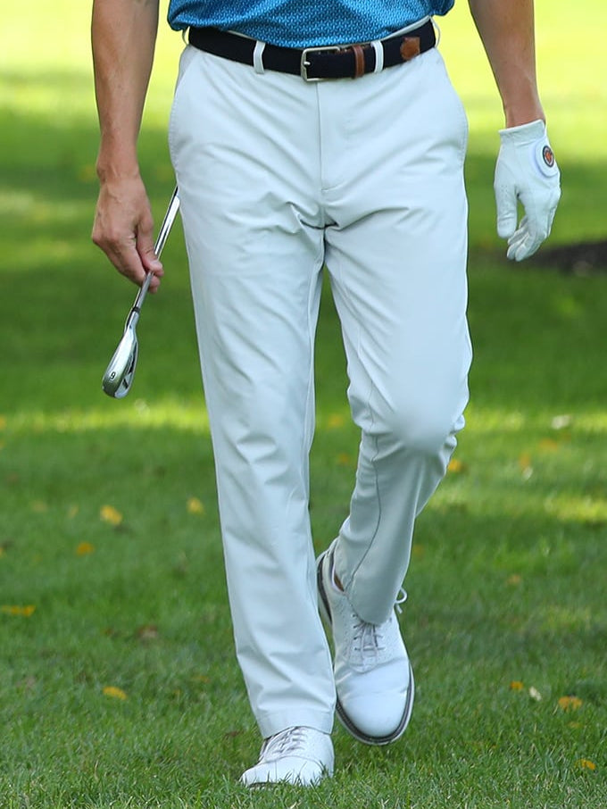 Two men walk on a lush golf course, each holding a club. They're wearing blue shirts and white pants, smiling and engaging in a lighthearted conversation