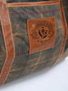 Close-up of a bag with brown leather accents and a plaid fabric. A leather patch reads "Donald Ross Sportswear" with a floral emblem, adding a vintage touch.