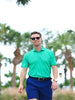 A man in a bright green polo and blue shorts walks confidently outdoors, wearing sunglasses