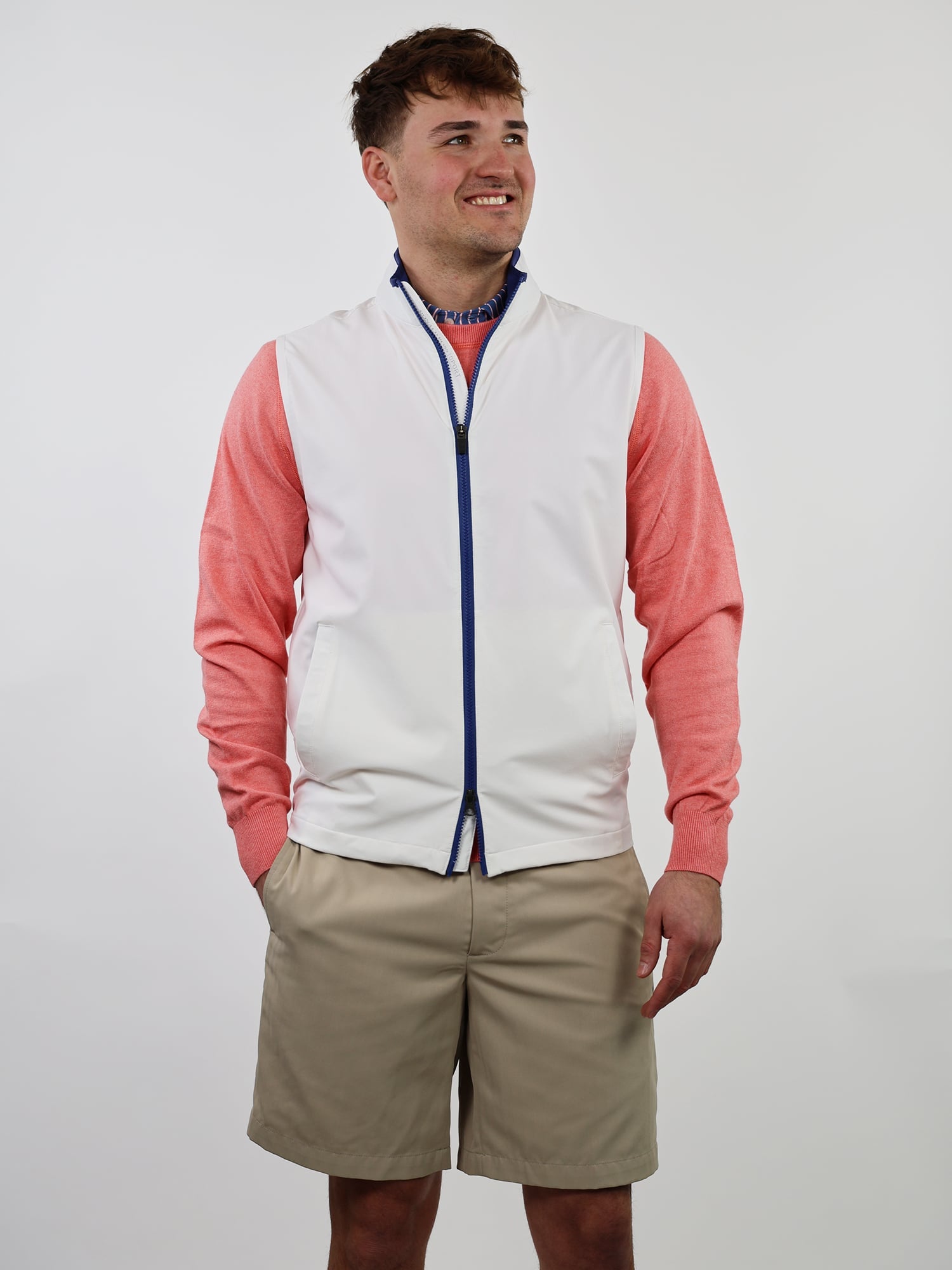 Man in a white zip-up vest over a coral long-sleeve shirt and beige shorts, smiling, with hands in pockets against a plain white background