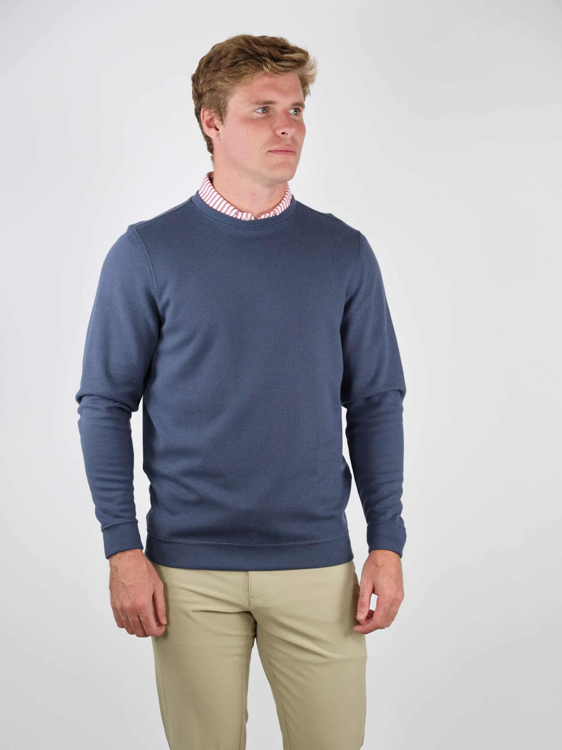 A man in a blue sweater and beige pants stands against a plain white background