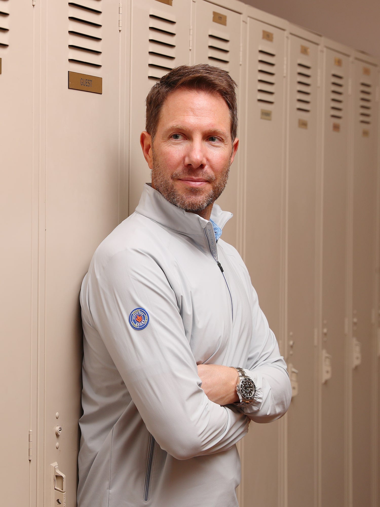 Man in a light gray jacket stands with arms crossed, leaning against beige lockers