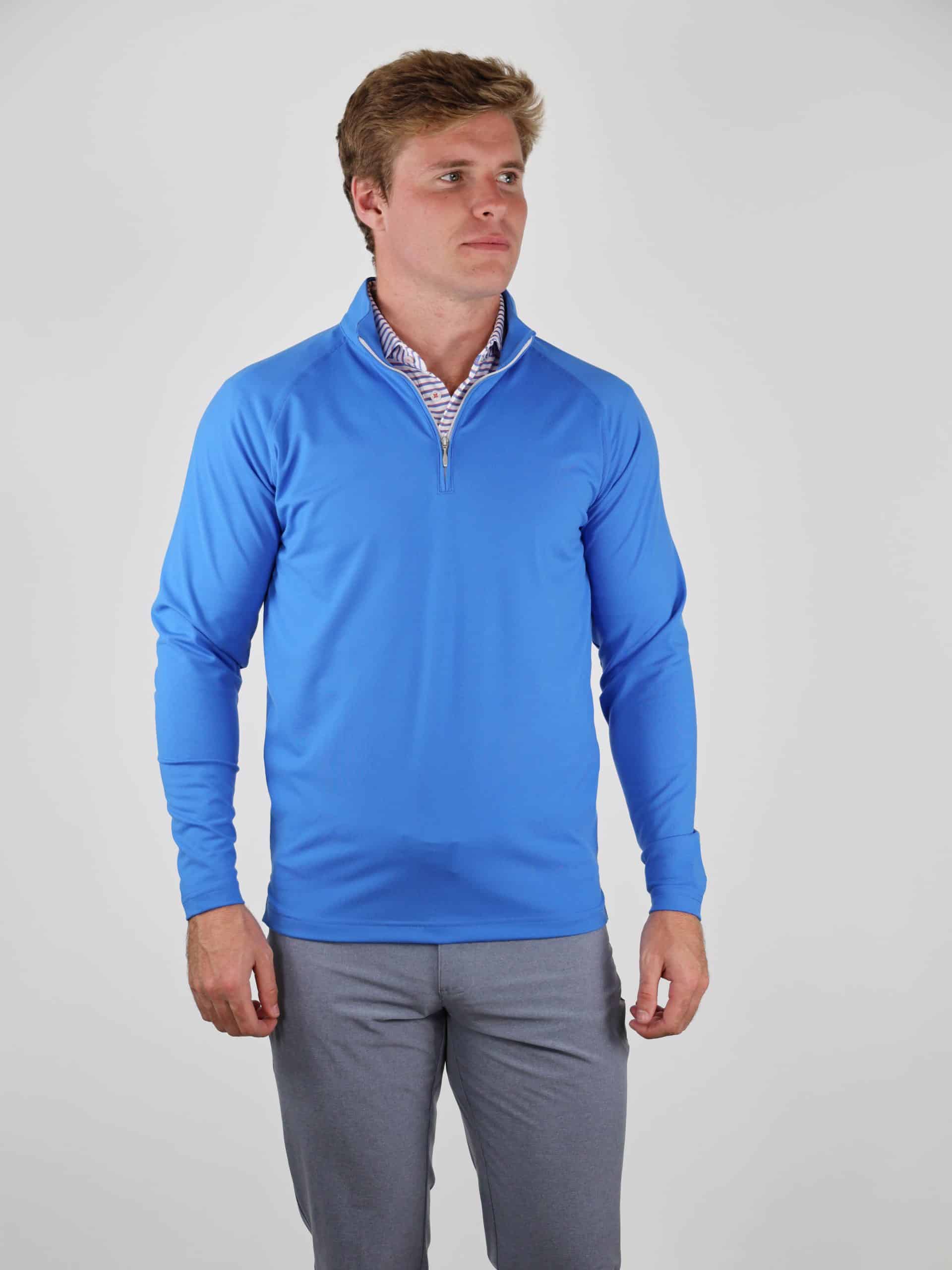 A person in a bright blue long-sleeve zip-up top and gray pants stands against a plain gray background, looking to the side with a neutral expression