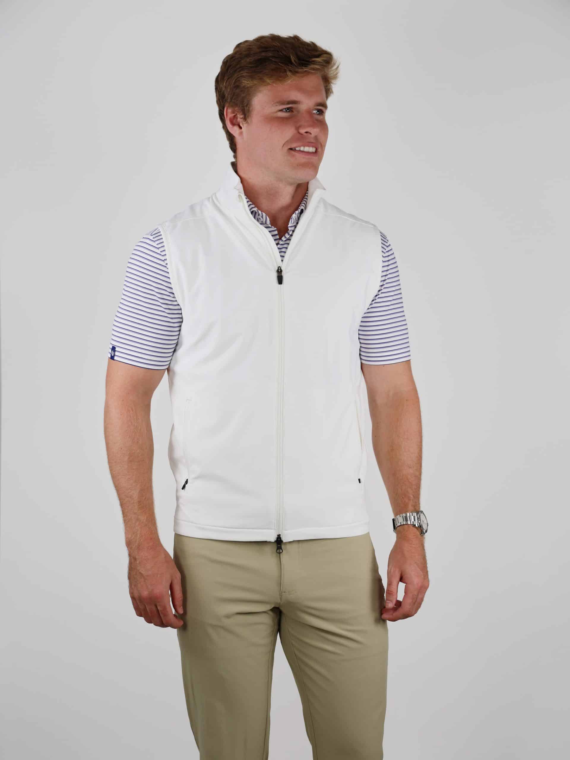 A man in a white zip-up vest and striped shirt smiles slightly, standing against a plain background. He wears khaki pants and a silver watch