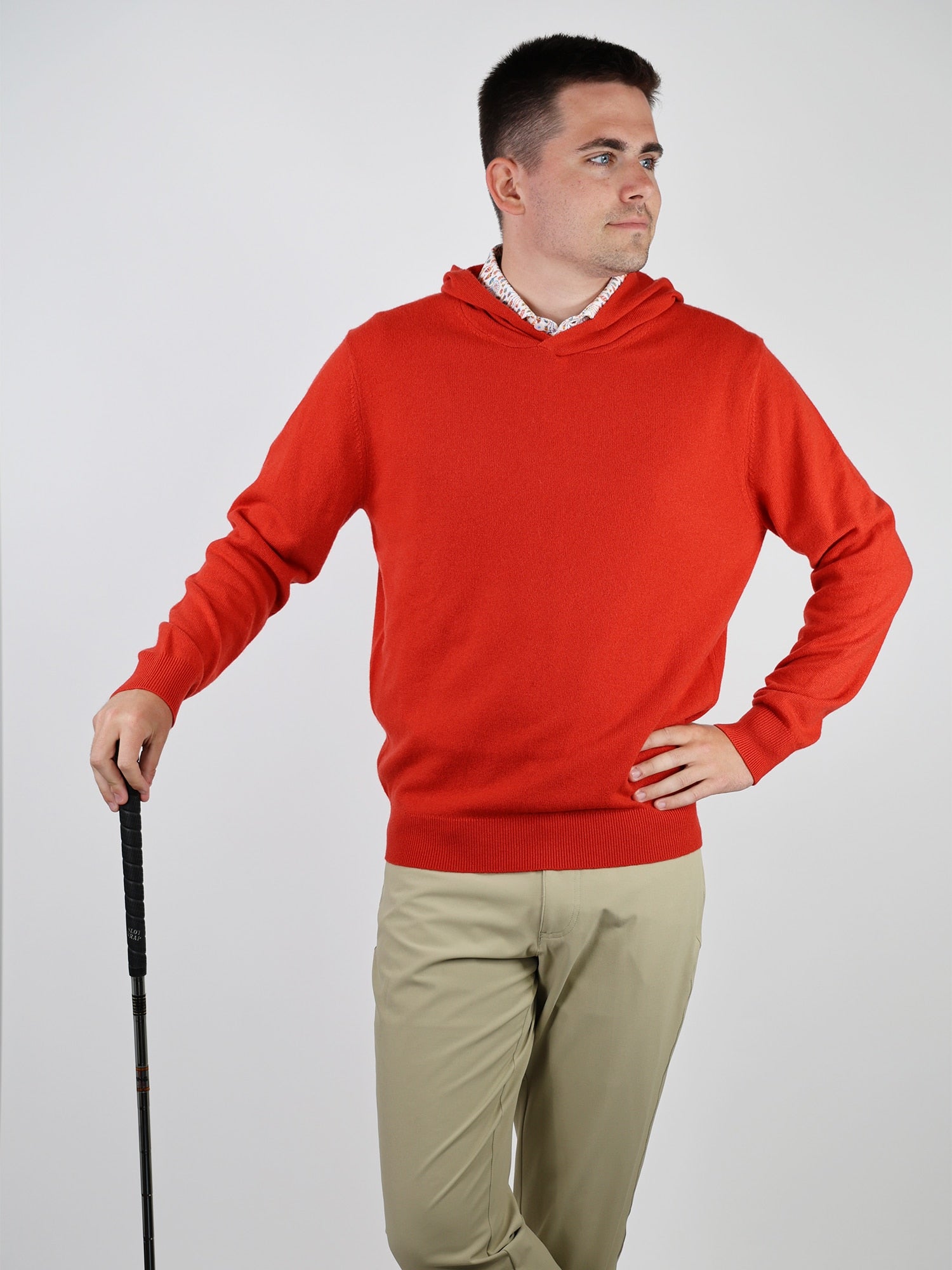 A person in a bright red sweater and beige pants holds a golf club and looks thoughtfully to the side