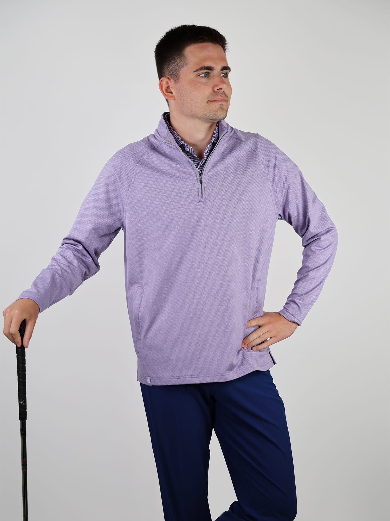 Man holding a golf club, wearing a light purple pullover and navy pants, stands confidently against a plain background