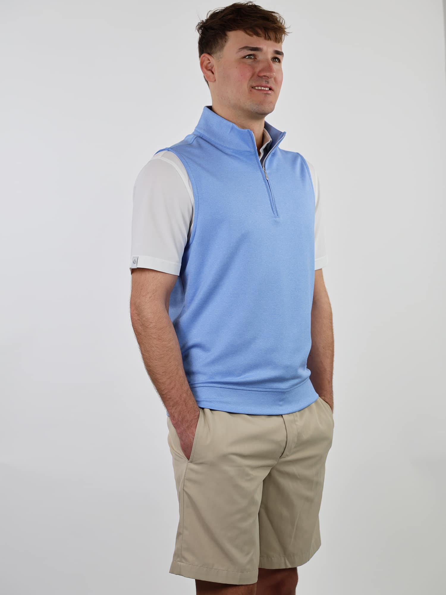A person wearing a light blue sleeveless pullover over a white shirt and khaki shorts stands with hands in pockets, looking to the side, smiling slightly