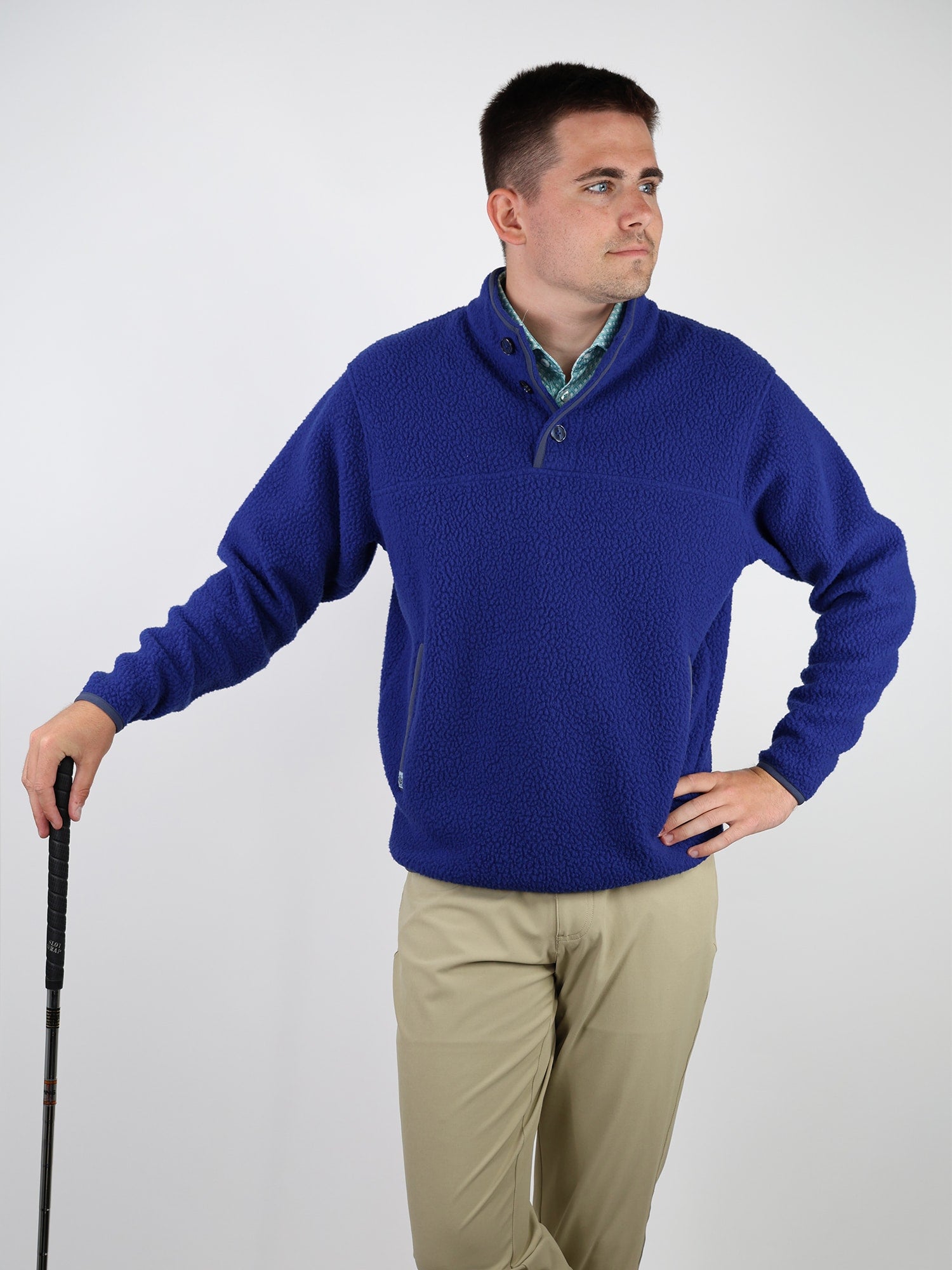 Man in a blue fleece sweater and beige pants holds a golf club, looking to the side with a thoughtful expression