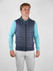 Man in a blue quilted vest and light blue long-sleeve shirt stands against a plain background, looking forward with a neutral expression