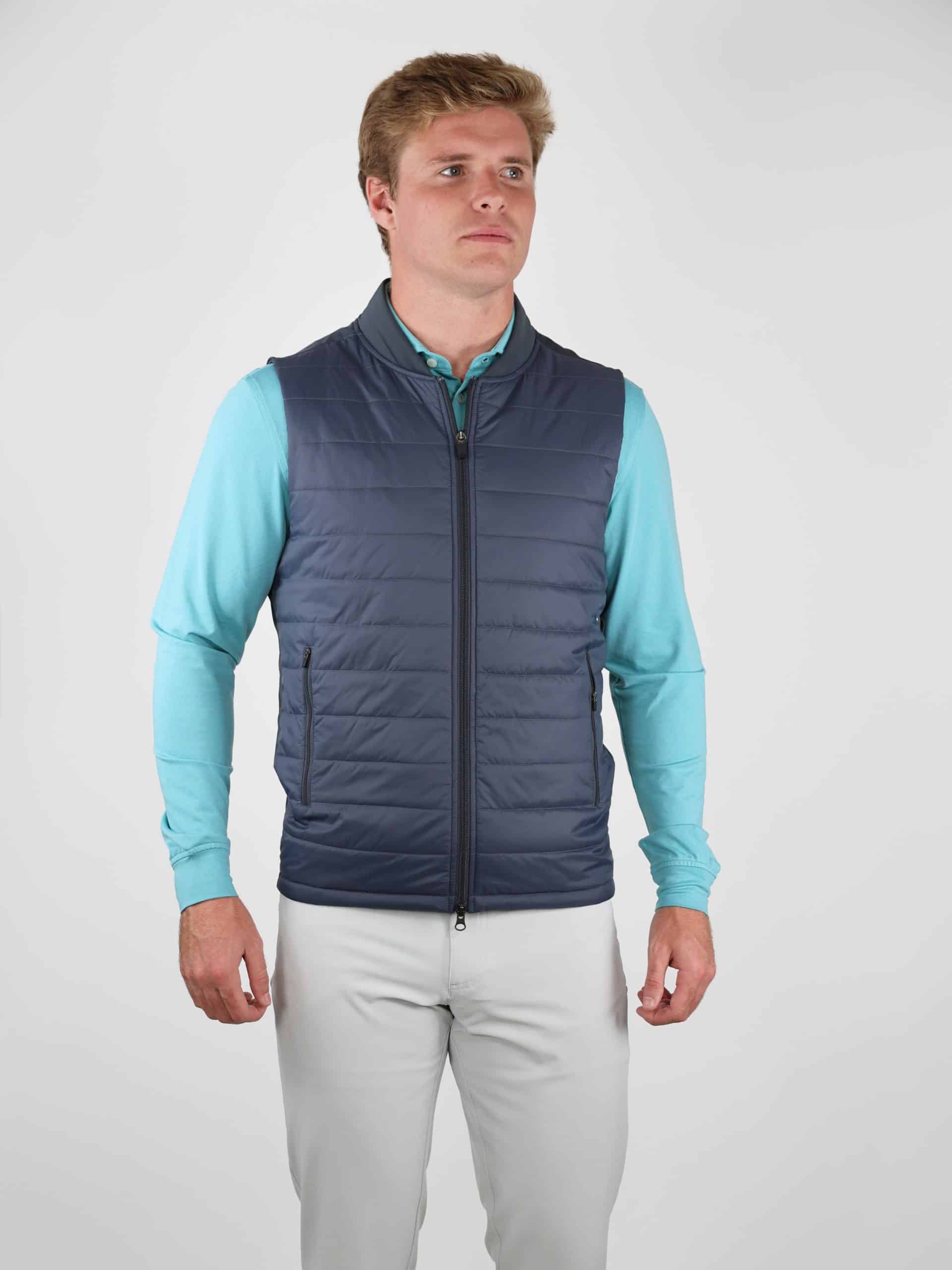 Man in a blue quilted vest and light blue long-sleeve shirt stands against a plain background, looking forward with a neutral expression