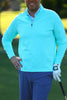 A man in a light blue pullover and gray pants stands with one hand on his hip and a golf club in his other hand, smiling against a blurred green background.