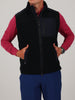 Person in a black zip-up fleece vest, red long-sleeve shirt, and blue pants, with hands in pockets. They wear a wristwatch, conveying a casual style.