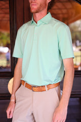 A man with a beard is standing in a light green striped polo shirt and beige pants, looking slightly to the side.